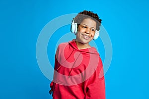 African-American girl in casual clothes listening to music in headphones against blue studio background. Happy, positive