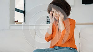 African American girl with curly hair woman sitting at home on couch touching massages temples feels headache dizziness