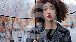 African American girl curly-haired sad woman in stylish coat stands in autumn on city street with transparent umbrella