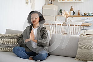African american girl doing yoga, meditation to keep calmness, sitting in lotus pose on sofa at home