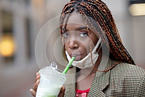 African American girl drinking matcha tea through the straw wearing a mace mask in the middle of coronavirus outbreak