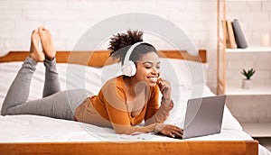 African American Girl In Headphones Using Laptop Lying On Bed