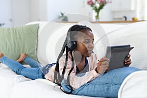 African american girl lying on couch, listening to music and using tablet at home, copy space