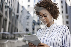 African-American girl standing on the street near the office building and holding a tablet. She uses on tablet