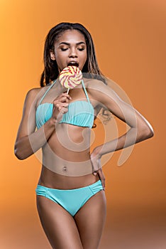 African american girl in swimwear eating sweet lollipop isolated on orange