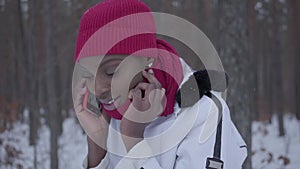 African american girl talking by cell phone standing in winter forest close up. Beautiful girl in white jacket turns and