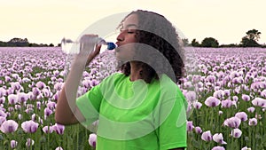 African American girl teenager female young woman walking drinking from a water bottle on path through field of pink poppy flowers