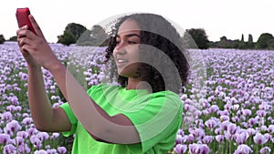 African American girl teenager female young woman walking through field of pink poppy flowers taking photographs on her smart phon