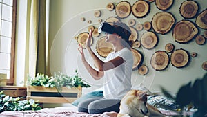 African American girl is using virtual reality glasses sitting on bed at home, she is moving hands and head and smiling