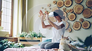 African American girl is using virtual reality glasses sitting on bed at home, she is moving hands and head and smiling