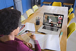 African american girl writing in notebook while teacher giving notes over video call on laptop