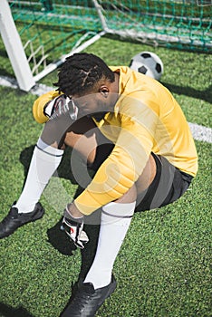 African american goalkeeper sitting at goalpost on pitch