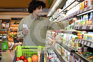 African American Guy Doing Grocery Shopping Holding Cellphone In Supermarket