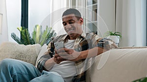 African American guy ethnic happy smiling man relaxing on comfortable couch sofa at home using mobile application