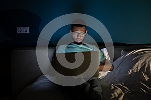 African American guy freelance lying in bed using laptop working remotely until late hours