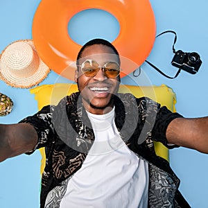 African American guy in funky outfit lying on lilo, taking selfie during beach vacation on blue background, top view