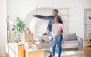 African American guy showing something to his happy girlfriend, standing among boxes in their new house, free space