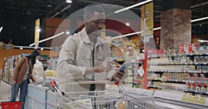 African American guy using smartphone app checking shopping list and looking at food products in cart in supermarket
