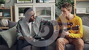 African American guy is using tablet to teach his Caucasian friend to play the guitar learning at home together. Men are