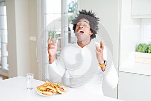 African American hungry man eating hamburger for lunch celebrating mad and crazy for success with arms raised and closed eyes