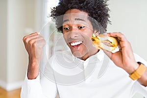 African American hungry man eating hamburger for lunch screaming proud and celebrating victory and success very excited, cheering