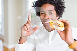 African American hungry man eating hamburger for lunch surprised with an idea or question pointing finger with happy face, number