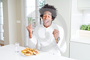 African American hungry man eating hamburger for lunch very happy and excited doing winner gesture with arms raised, smiling and