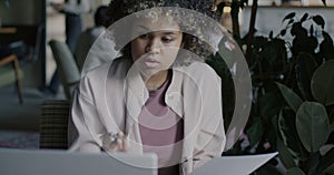 African American lady entrepreneur working in cafe reading business document and looking at laptop screen