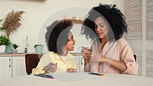 African American litle girl draws with felttip pens in album. Mother and daughter are posing sitting at table in bright