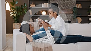 African American loving mother mommy lying on sofa relaxing at home living room with little daughter child talking