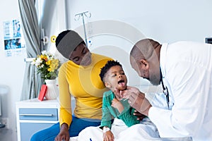 African american male doctor examining mouth of boy patient with mother in hospital