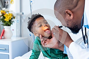 African american male doctor examining mouth of boy patient with penlight in hospital