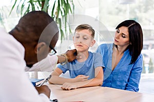 African-American male doctor in his modern office clinic examining throat of little boy with pretty mother watching.