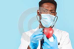 African american male doctor with stethoscope in mask holding red heart on blue background. cardiologist