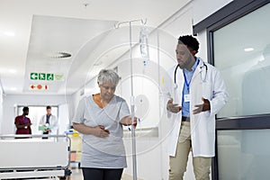 African american male doctor talking to diverse senior female patient holding drip stand in corridor