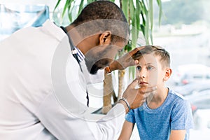 African American male doctor testing patient`s eye of a little boy in clinic. Medical examination concept