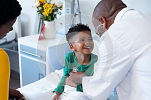 African american male doctor using stethoscope on smiling boy patient in hospital