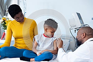 African american male doctor vaccinating boy patient, with mother in hospital