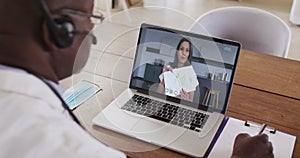 African american male doctor wearing phone headset taking notes while having a video call on laptop