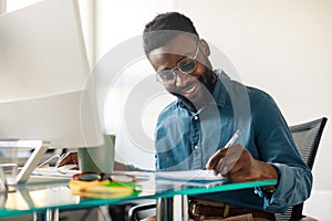 African american male entrepreneur working on modern computer and taking notes, sitting at workpace in office interior