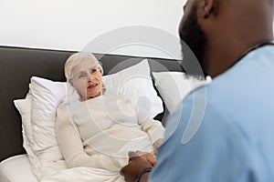 African american male health worker supporting caucasian senior woman lying on the bed at home