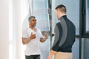 African American male office worker having very serious conversation with Caucasian colleague standing by window and
