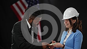 African American male politician passing cash money to Caucasian woman in hard hat in camera flashes. Confident man
