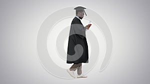 African american male student in graduation robe texting on the phone while walking with his diploma on gradient