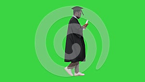 African american male student in graduation robe texting on the phone while walking with his diploma on a Green Screen