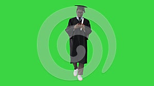 African american male student in graduation robe texting on the phone while walking with his diploma on a Green Screen