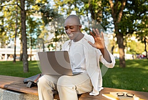 African american male student with laptop attending online lecture, waving hand at webcamera, sitting outdoors