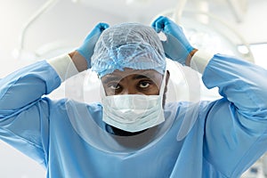 African american male surgeon in gown and cap tying on mask in operating theatre