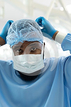 African american male surgeon in gown and cap tying on mask in operating theatre, copy space