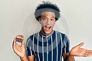 African american man with afro hair holding cake slice celebrating achievement with happy smile and winner expression with raised